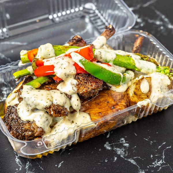 Baked potato topped with steak, red and green bell peppers, and creamy sauce in a clear container.