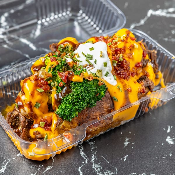 A loaded baked potato with cheese, broccoli, bacon, chives, and sour cream in a plastic container.